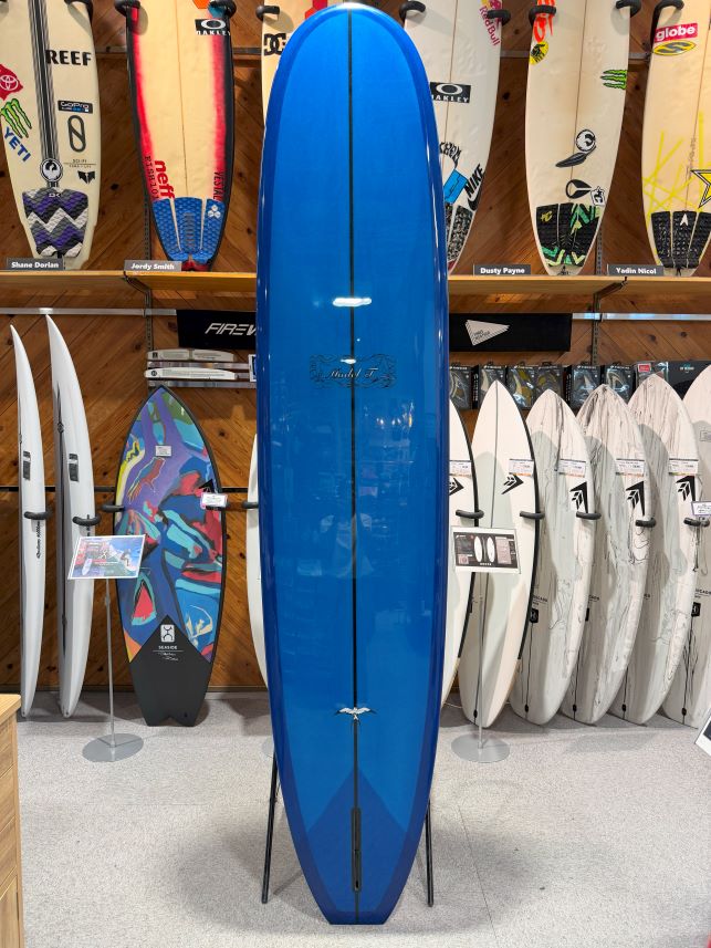IMG_0281 - SHOP NEWS | ムラサキスポーツオンラインストア 通販 Blue surfboard on a stand in a shop, surrounded by many other surfboards on racks and walls.