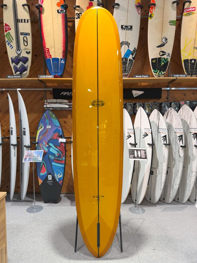 IMG_0289 - SHOP NEWS | ムラサキスポーツオンラインストア 通販 Bright yellow longboard on a display stand in a surf shop, with other boards lining the walls behind it and racks in the foreground.