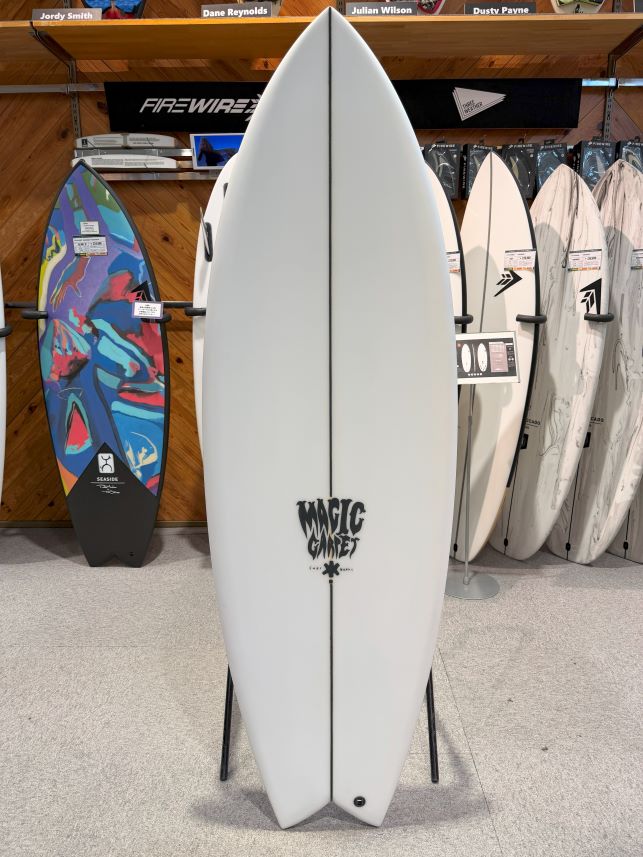 White surfboard standing upright on a display stand in a surf shop, with other boards on racks in the background.