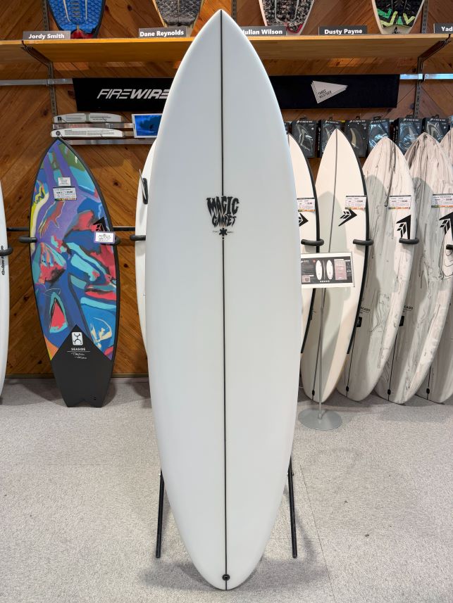 Tall white surfboard on a display stand in a surf shop, with colorful and white boards on racks behind it.