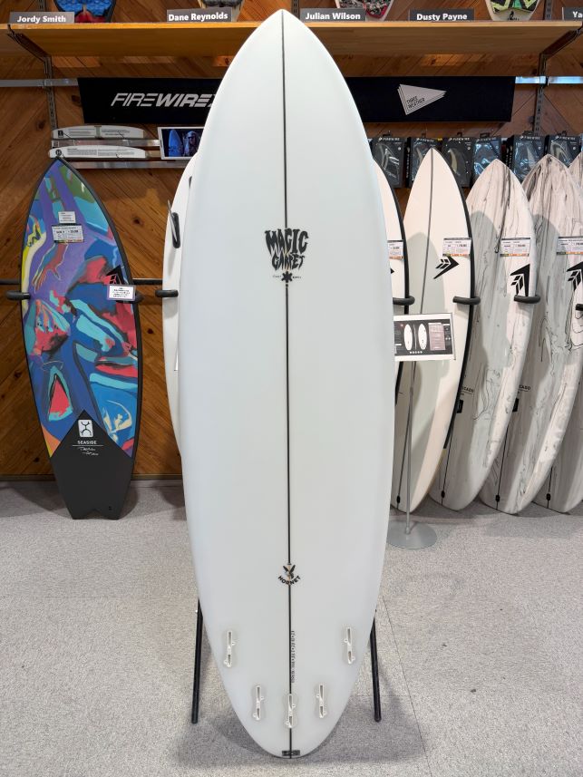 White surfboard on a display stand in a shop, with a vertical center stringer and 'Magic Corner' logo; other surfboards and racks are visible in the background.