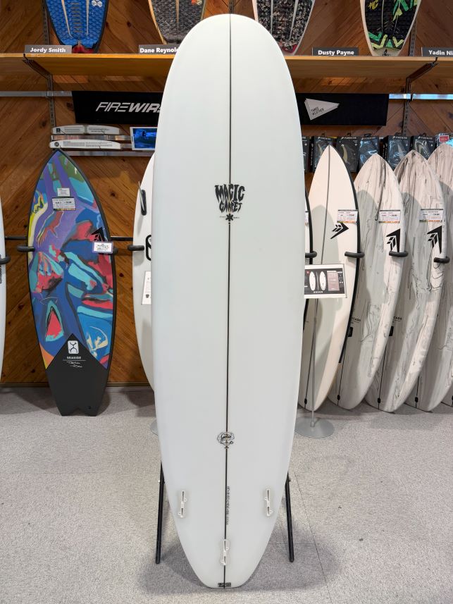 White surfboard on a display stand in a shop, with other boards and racks in the background and the word 'Magic' near the center.