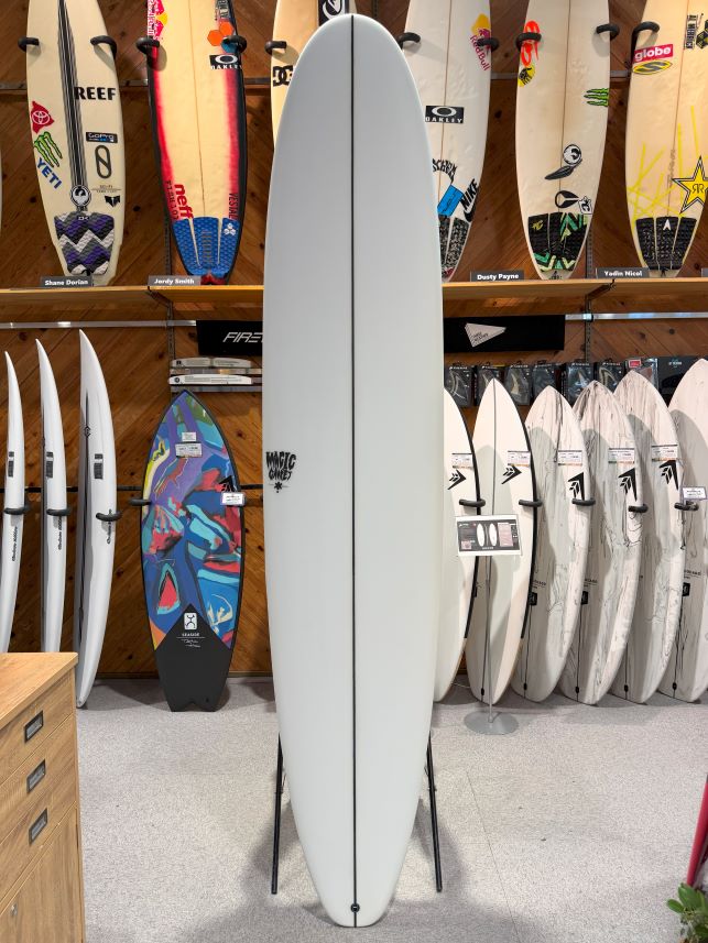 Tall light-gray longboard on a stand in a surf shop, surrounded by colorful boards on racks in the background.