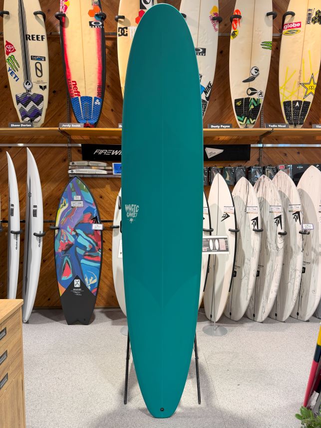 Teal longboard standing upright on a stand in a surf shop, surrounded by white and colorful boards on racks.