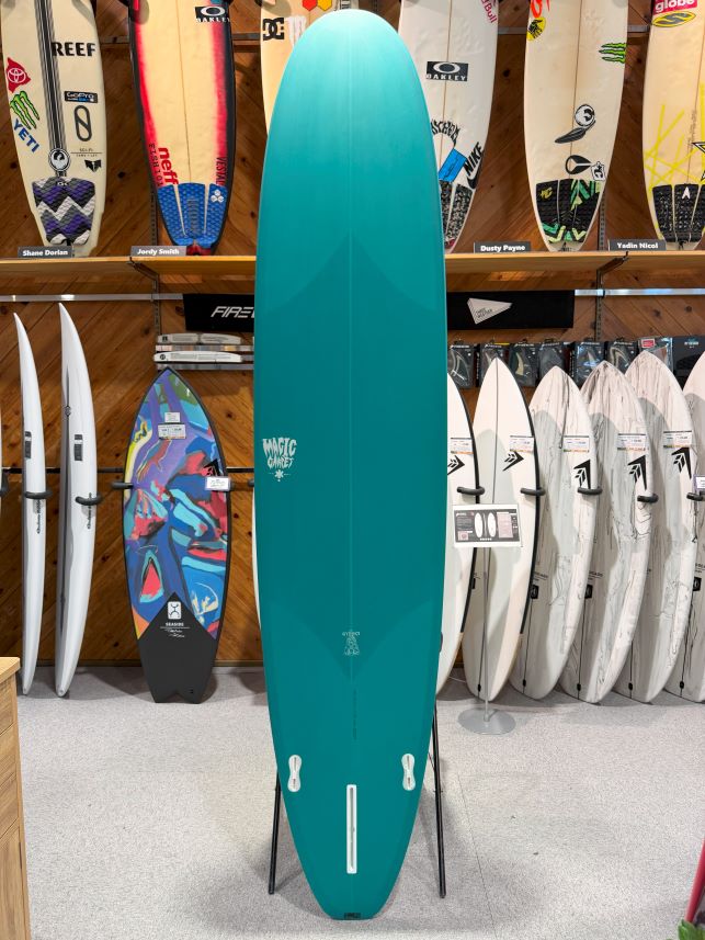 Teal surfboard on a stand in a surf shop, surrounded by a variety of boards on racks and walls behind it.