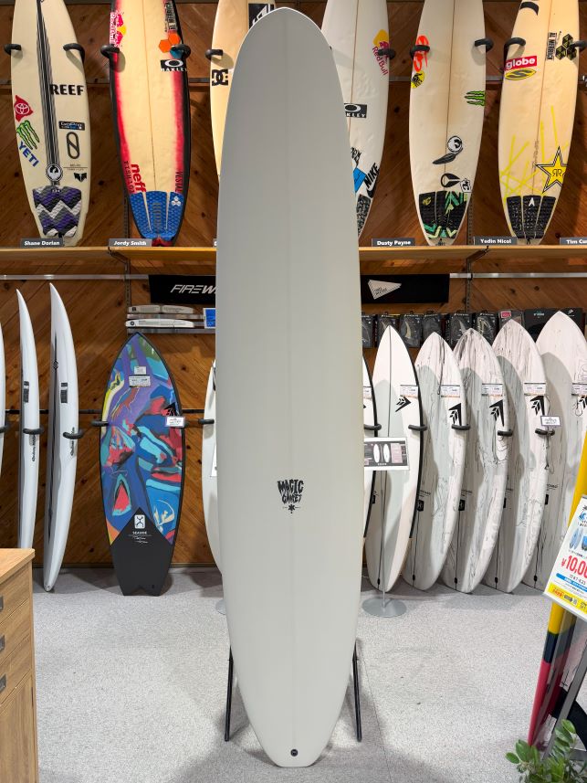 Large pale gray surfboard on a stand in a showroom, with numerous colorful boards displayed on wooden racks in the background.