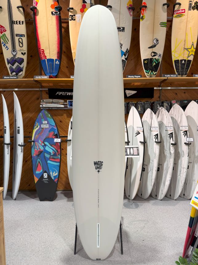 Tall white surfboard standing upright on a display rack in a surf shop, with colorful boards on shelves behind it.