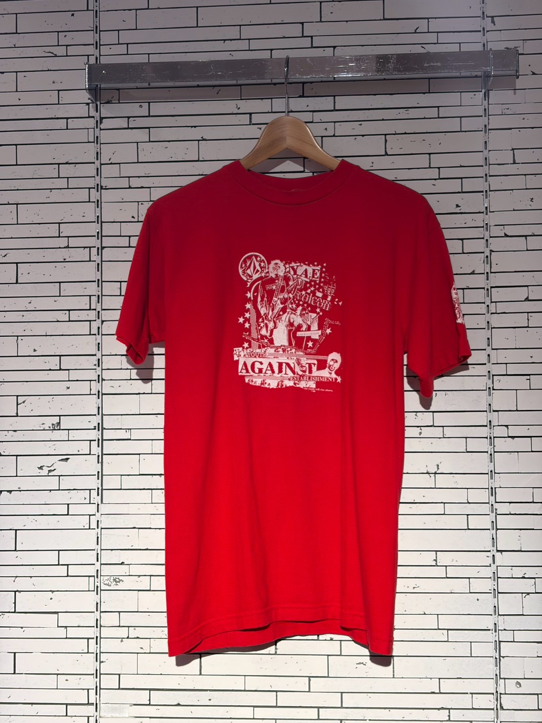 Red short-sleeve T-shirt on a wooden hanger against a white brick wall, featuring a white graphic with the word 'AGAINST'.