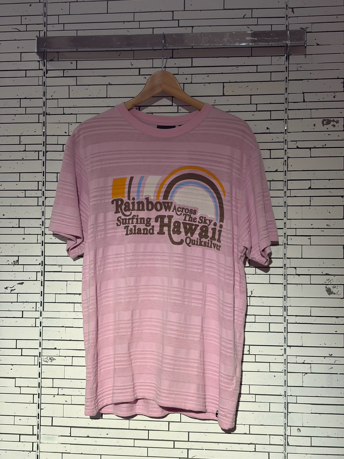 Pink T-shirt on a wooden hanger with a retro rainbow graphic and brown text reading 'Hawaii' and 'Rainbow Across The Sky Surfing Island Quiksilver' (informative description)