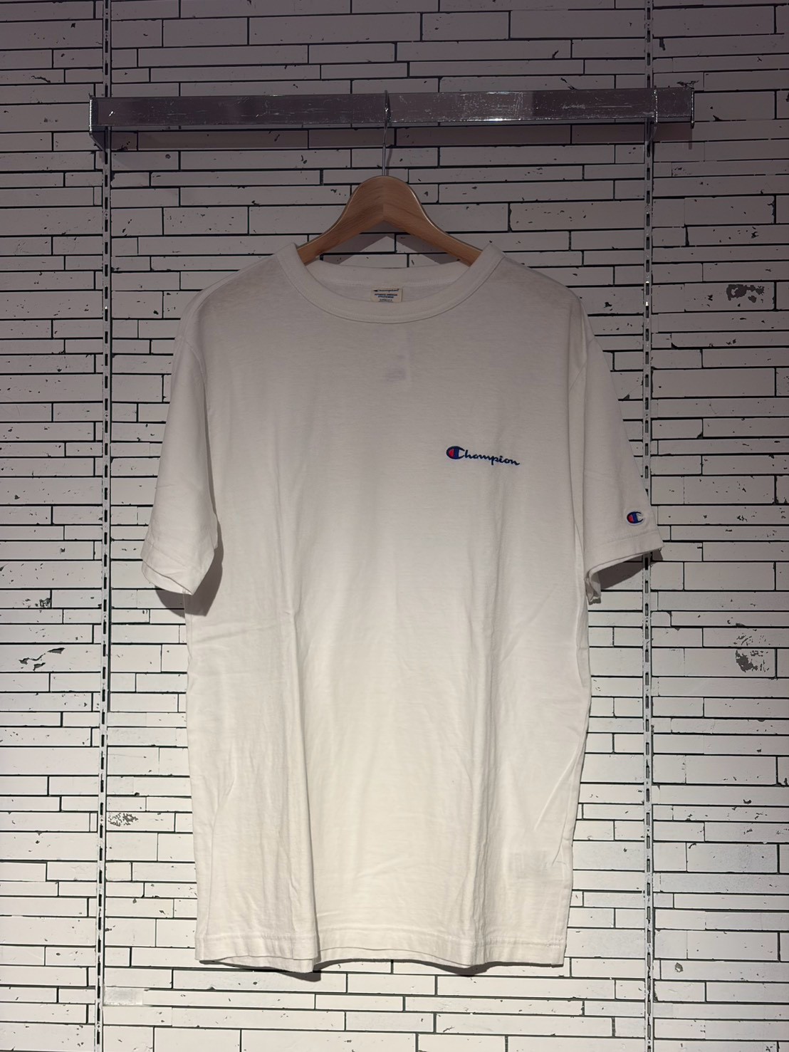 White Champion t-shirt on a wooden hanger against a white brick wall, showing a small Champion logo on the chest.