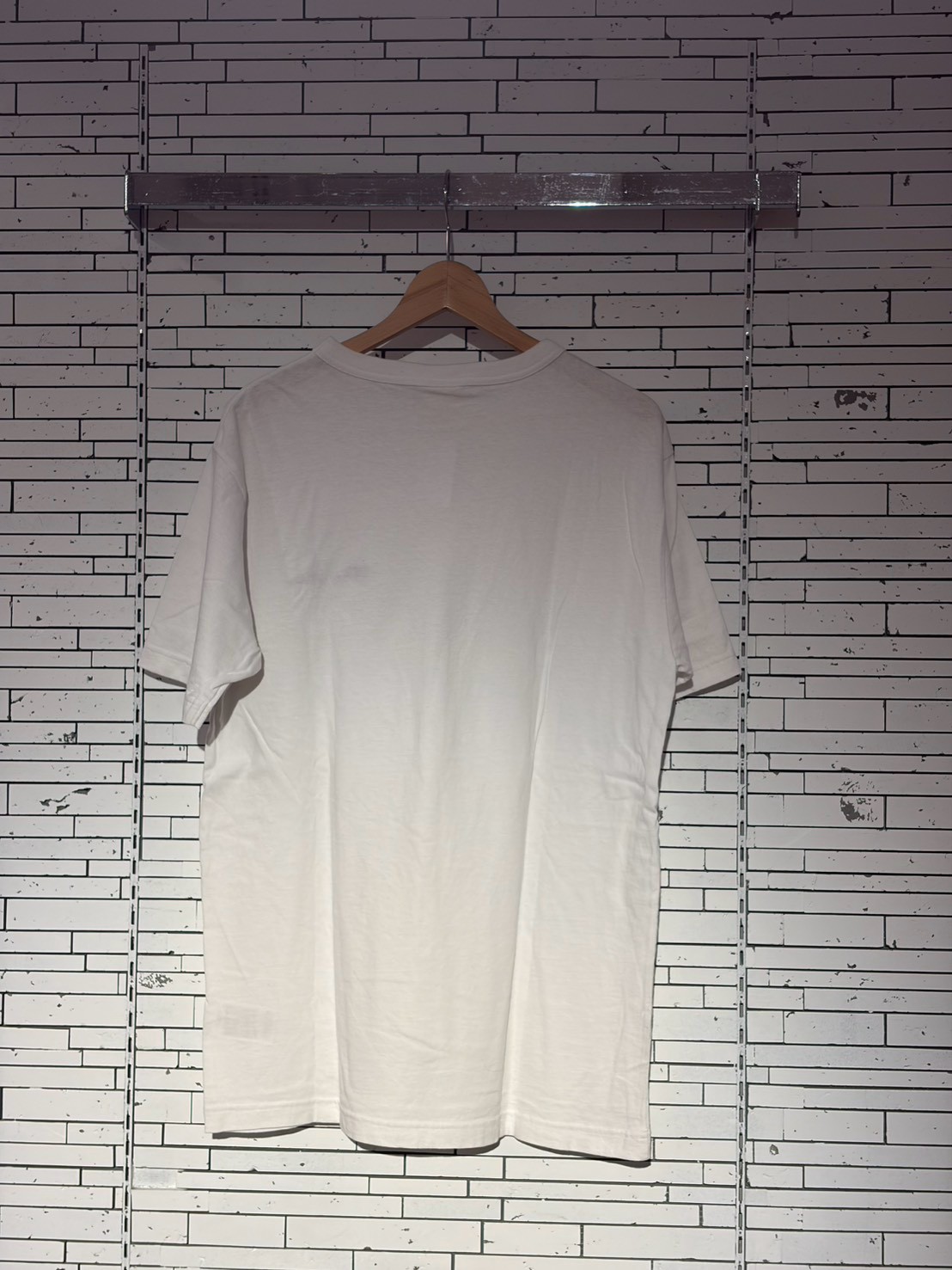 White t-shirt on a wooden hanger against a brick wall with a metal rack above.