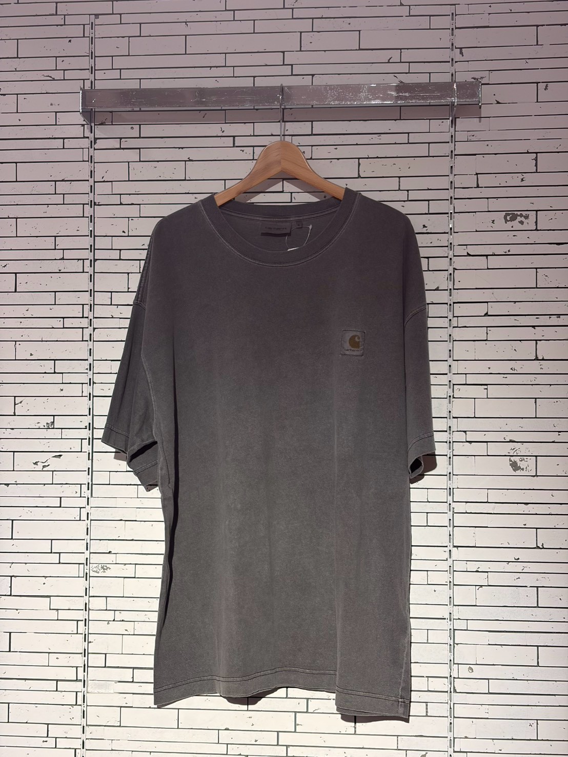 Gray oversized short-sleeve t-shirt on a wooden hanger against a white brick wall; small Carhartt logo on the left chest pocket.