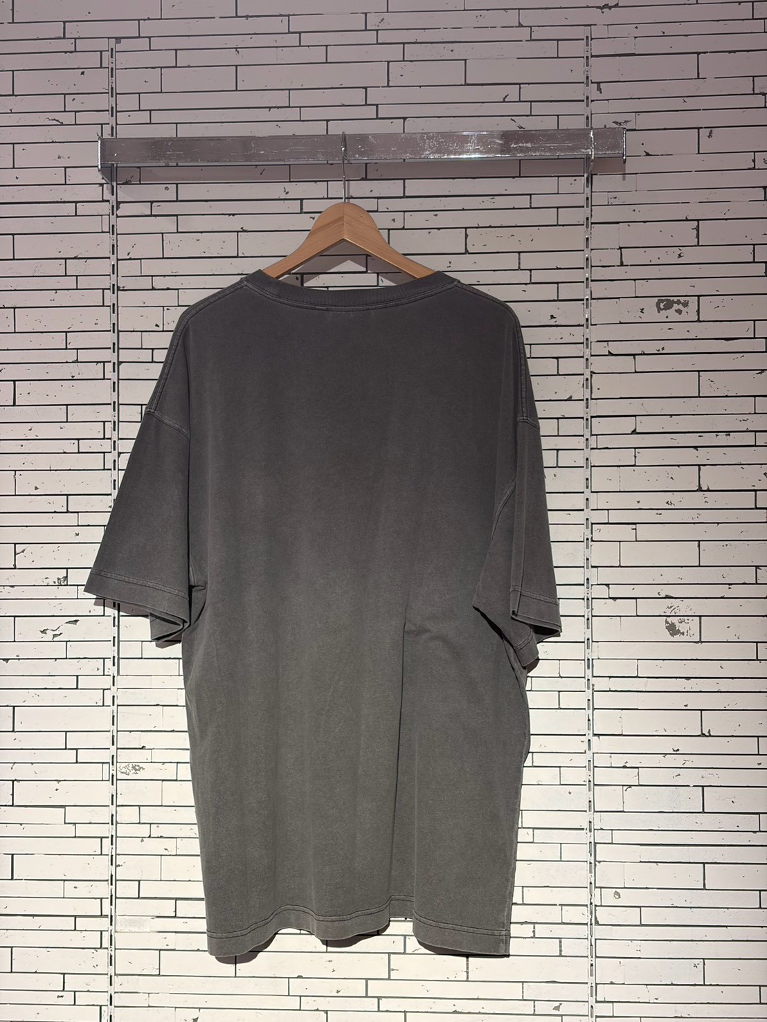 Gray oversized T-shirt on a wooden hanger against a white tiled wall.