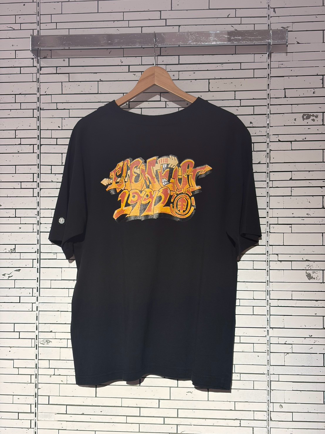Black T-shirt on a wooden hanger with an orange graffiti-style graphic text on the chest, against a white brick wall backdrop.