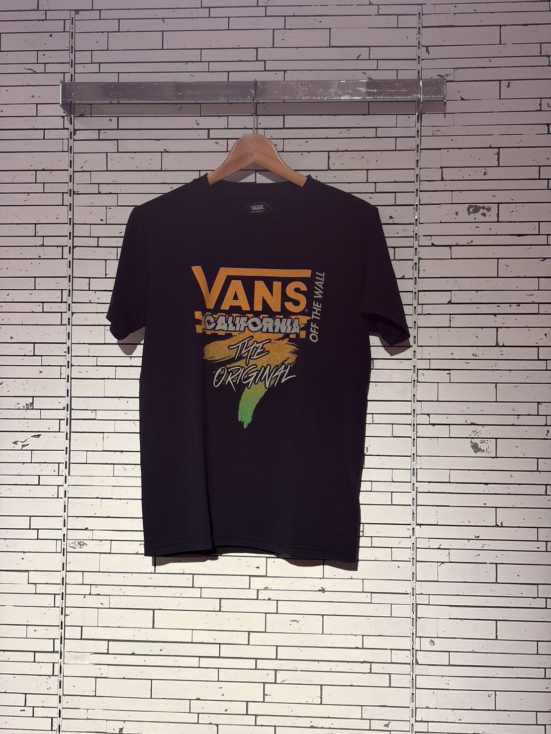 Black Vans T-shirt on hanger against a white brick wall; graphic reads 'VANS CALIFORNIA The ORIGINAL' with colorful brush strokes and 'OFF THE WALL' text along the side.