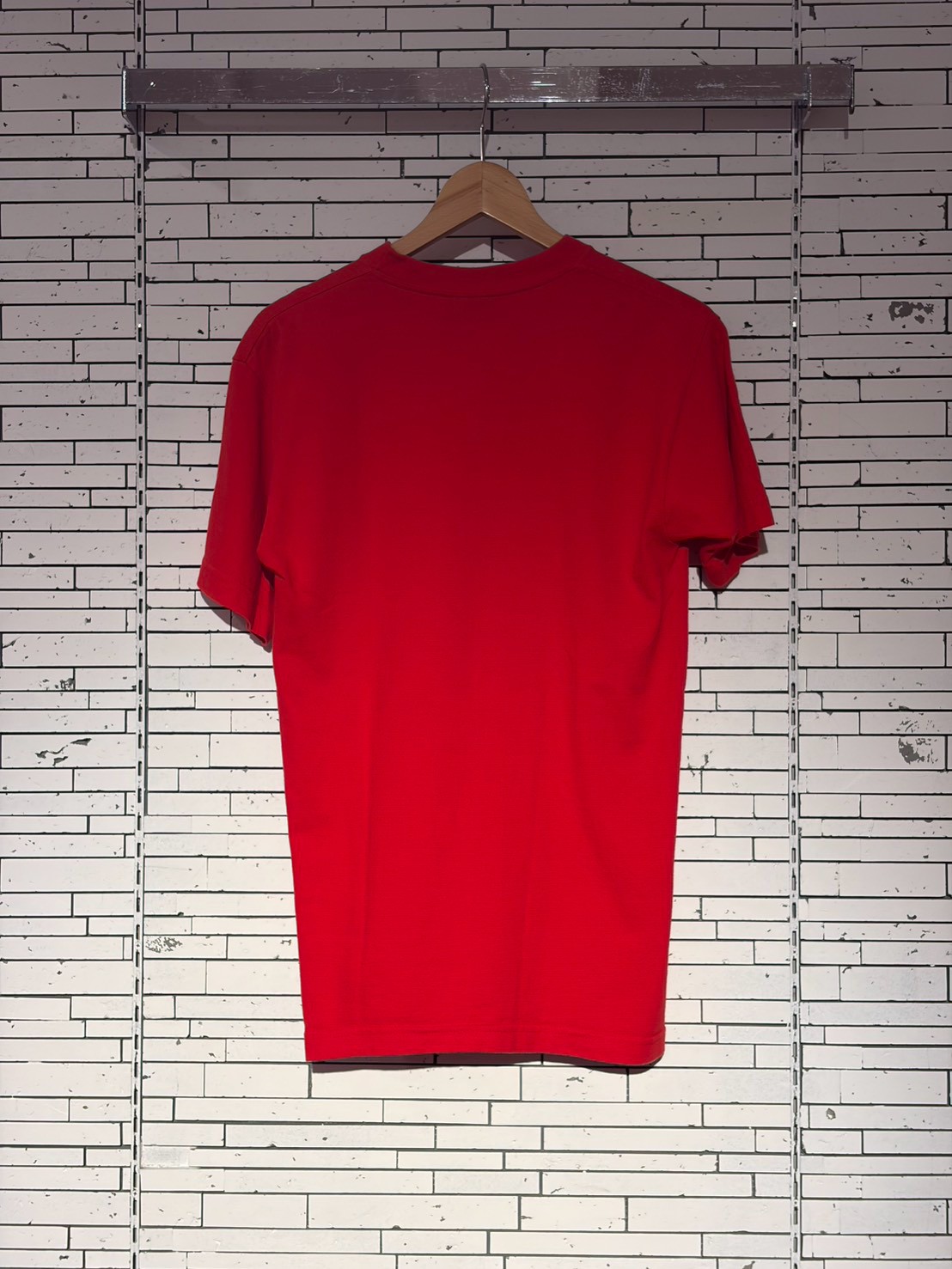 Red short-sleeve T-shirt on a wooden hanger against a light brick wall.