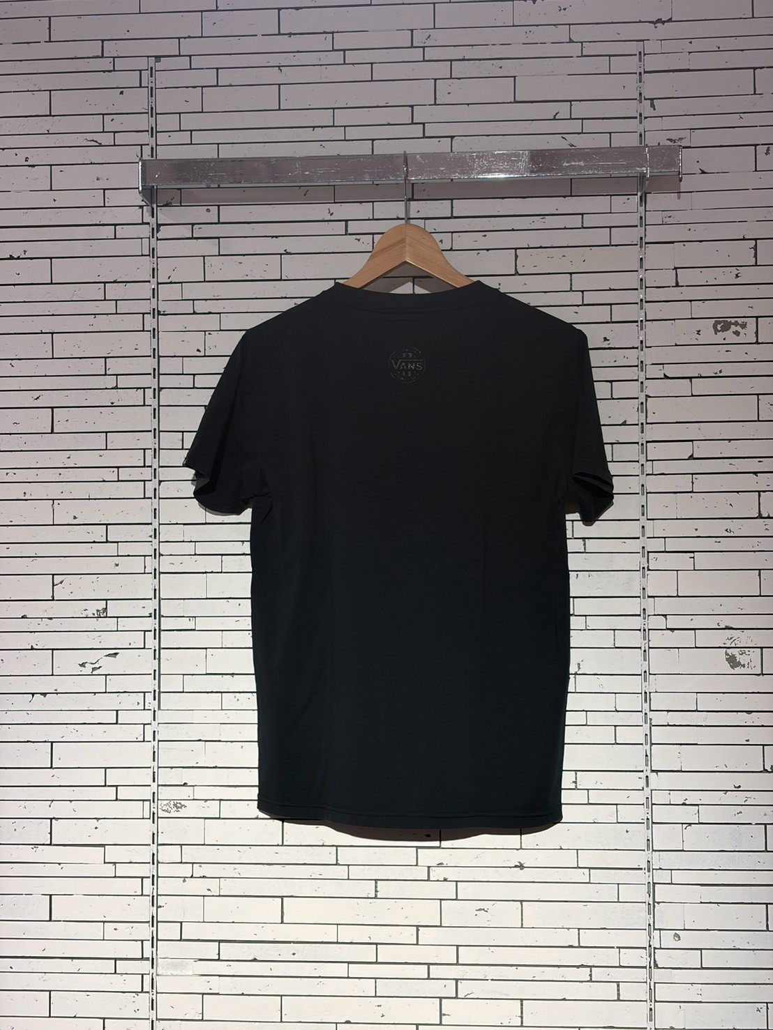 Black T-shirt on a wooden hanger against a white brick wall; small Vans logo on the back near the neckline.