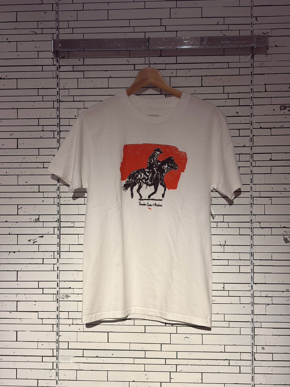 White t-shirt on a wooden hanger against a tiled wall; graphic of a rider on a horse over an orange-red brush stroke with small text beneath