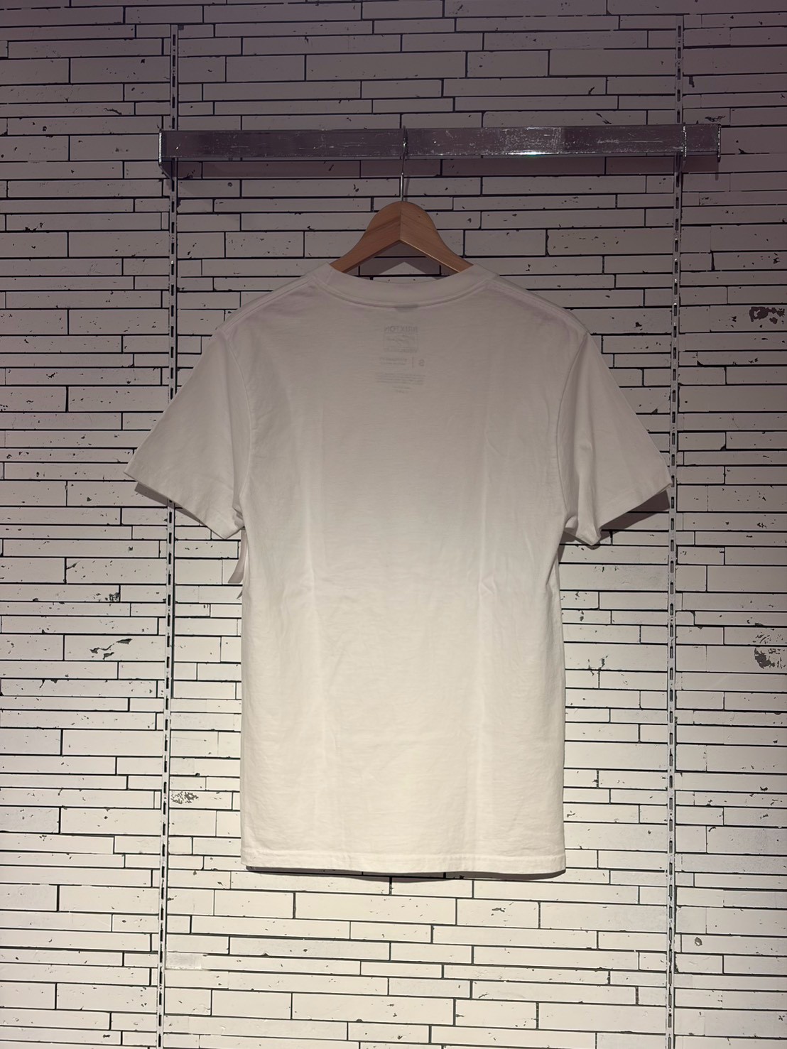White short-sleeve T-shirt on a wooden hanger against a distressed white brick wall, displayed for sale.