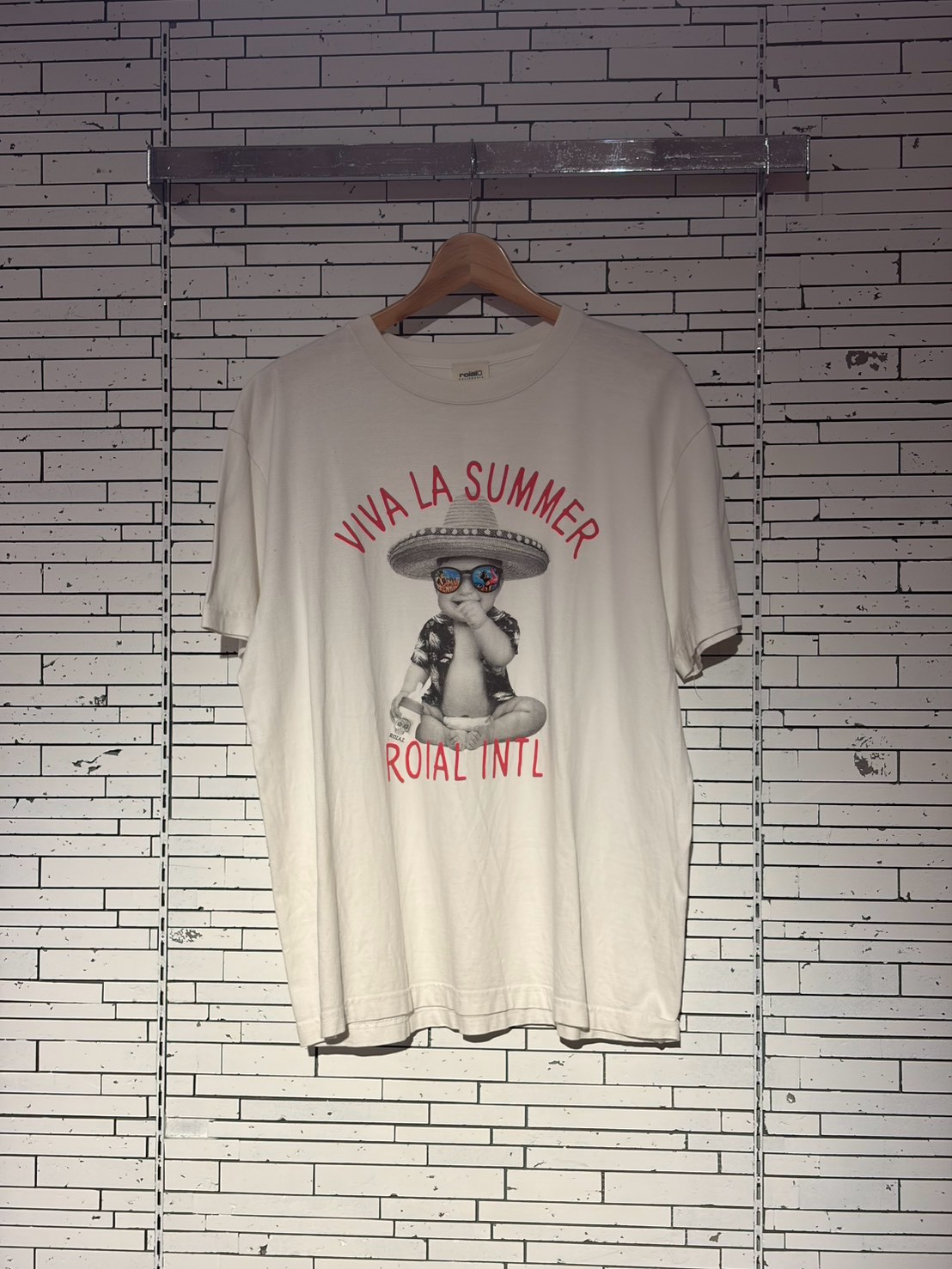 White T-shirt on a wooden hanger hanging against a white tiled wall, featuring a graphic of a baby wearing a sombrero and colorful sunglasses with the text 'VIVA LA SUMMER' and 'ROIAL INTL'