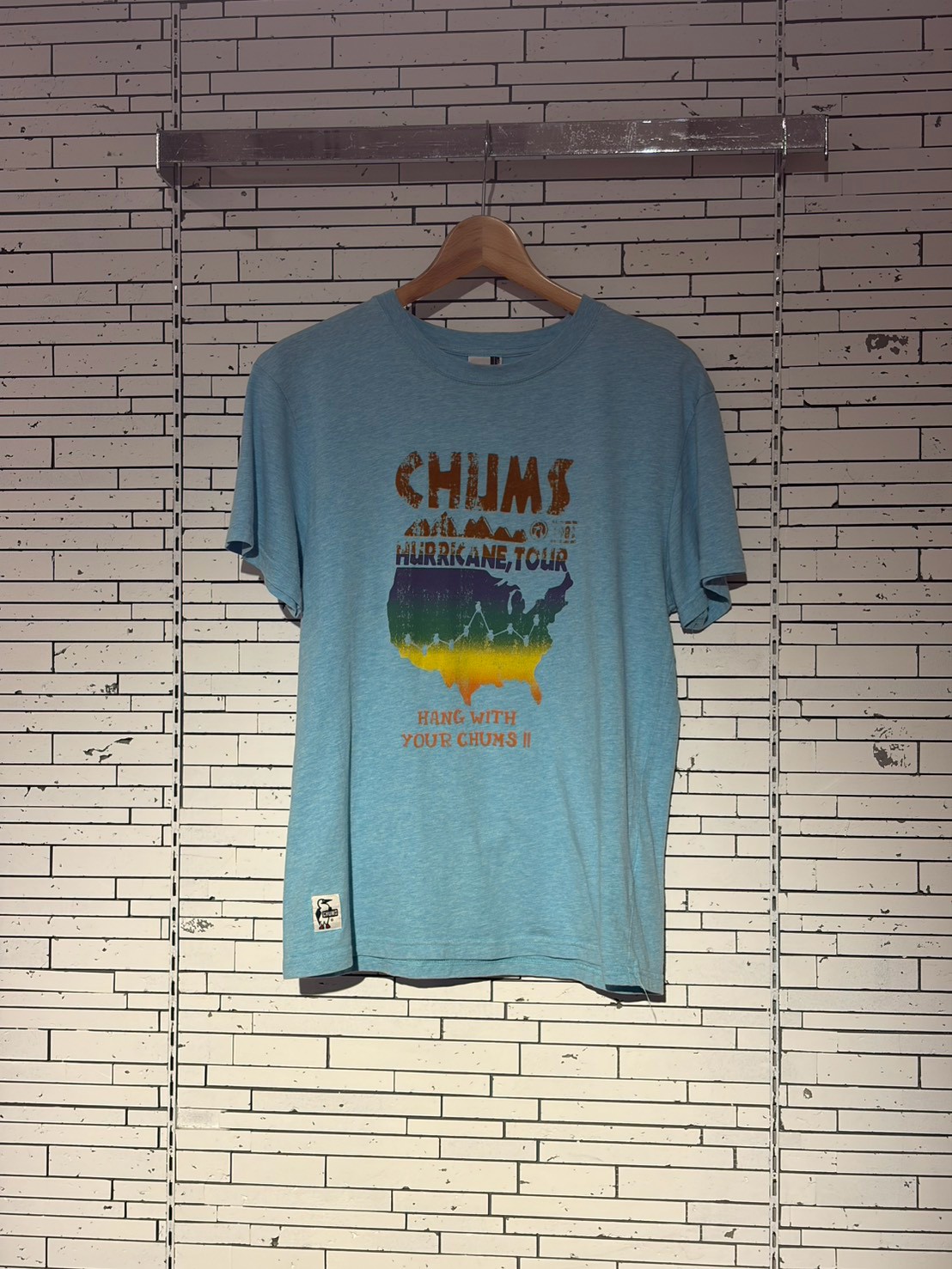 Light blue CHUMS t-shirt on a wooden hanger displaying the Hurricane Tour graphic with a rainbow US map and the caption 'Hang with your chums II'.