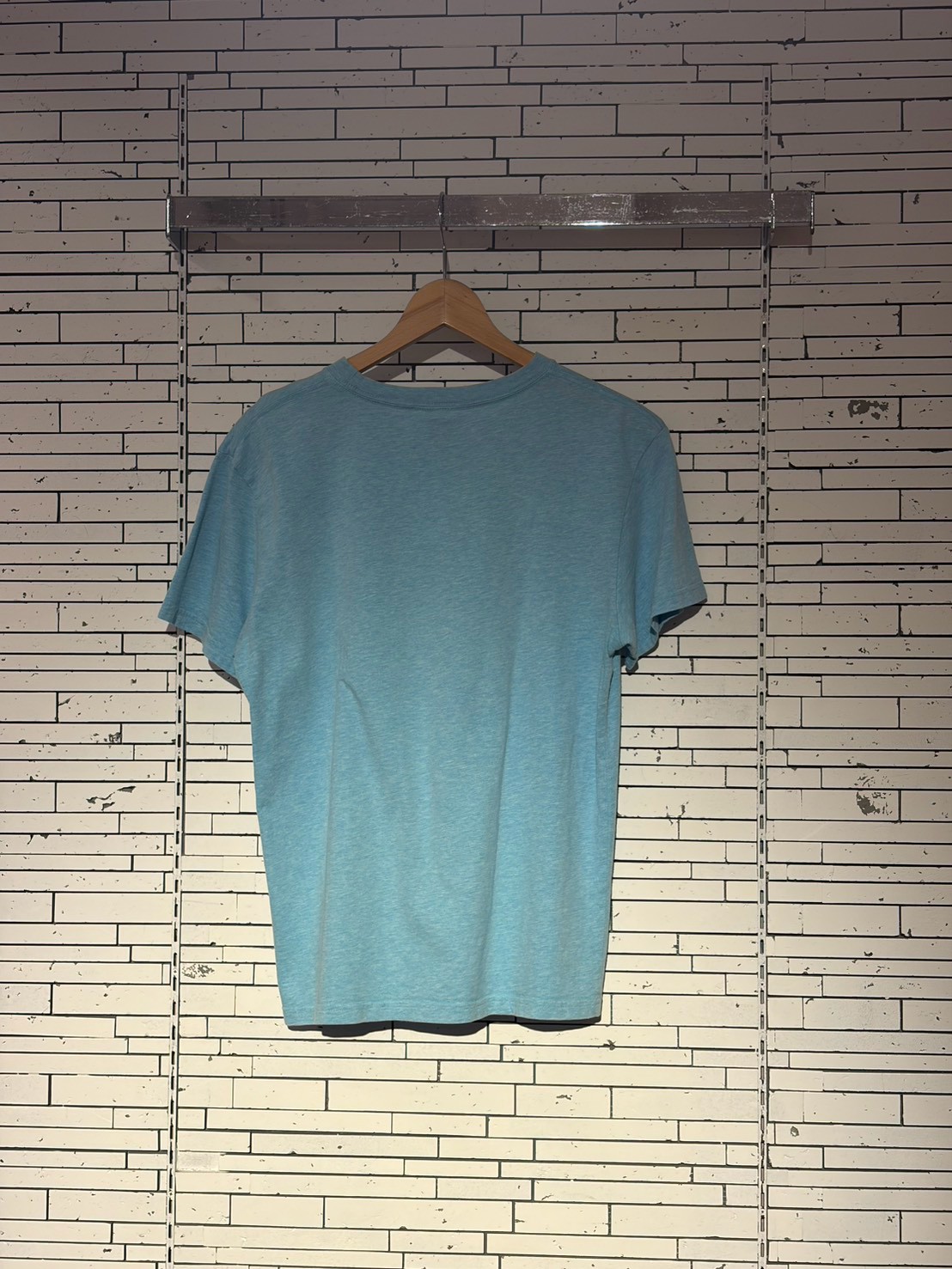 Light blue short-sleeve T-shirt on a wooden hanger against a pale, striped wall backdrop.”