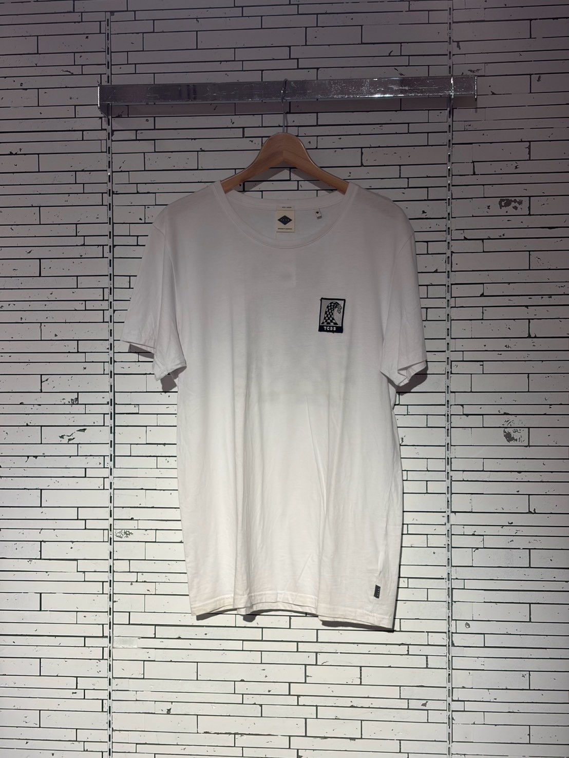 White t-shirt on a wooden hanger against a white brick wall, small square logo on the left chest area.