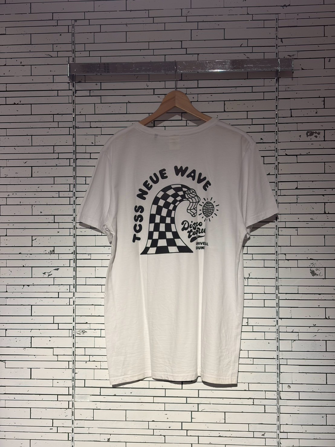 White T-shirt on a hanger with a black graphic: a large checkered wave motif, plus small text and a disco ball illustration nearby.