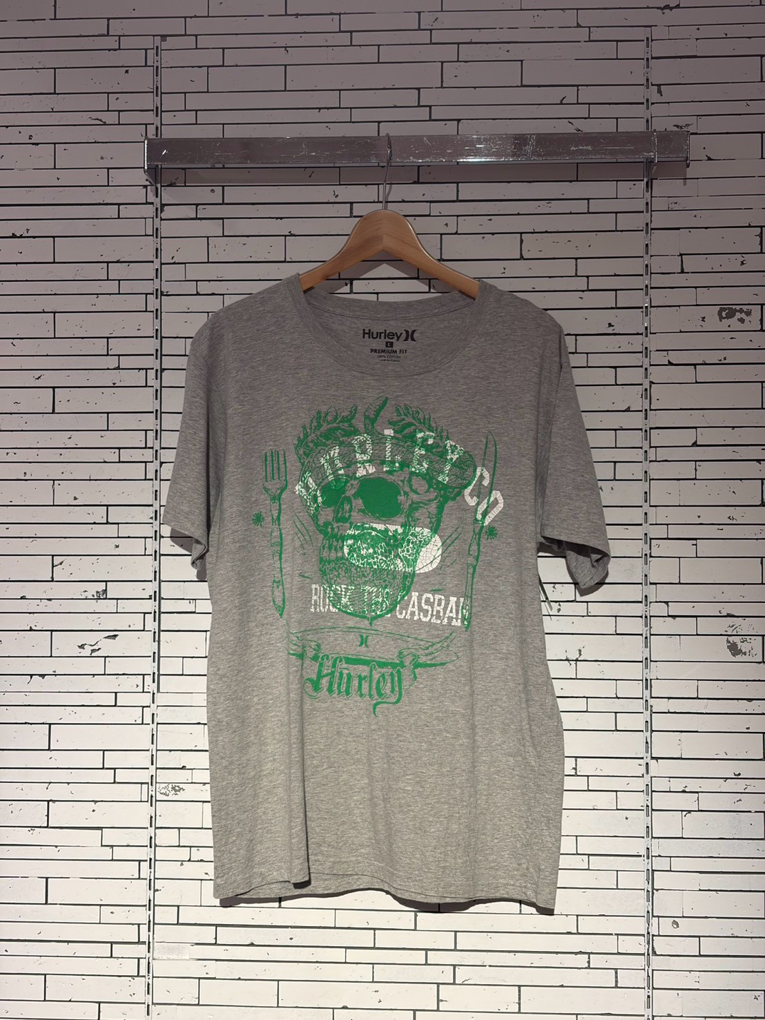 Gray Hurley Premium Fit T-shirt on a wooden hanger with a green skull graphic, hung against a white brick wall framed by metal rails.