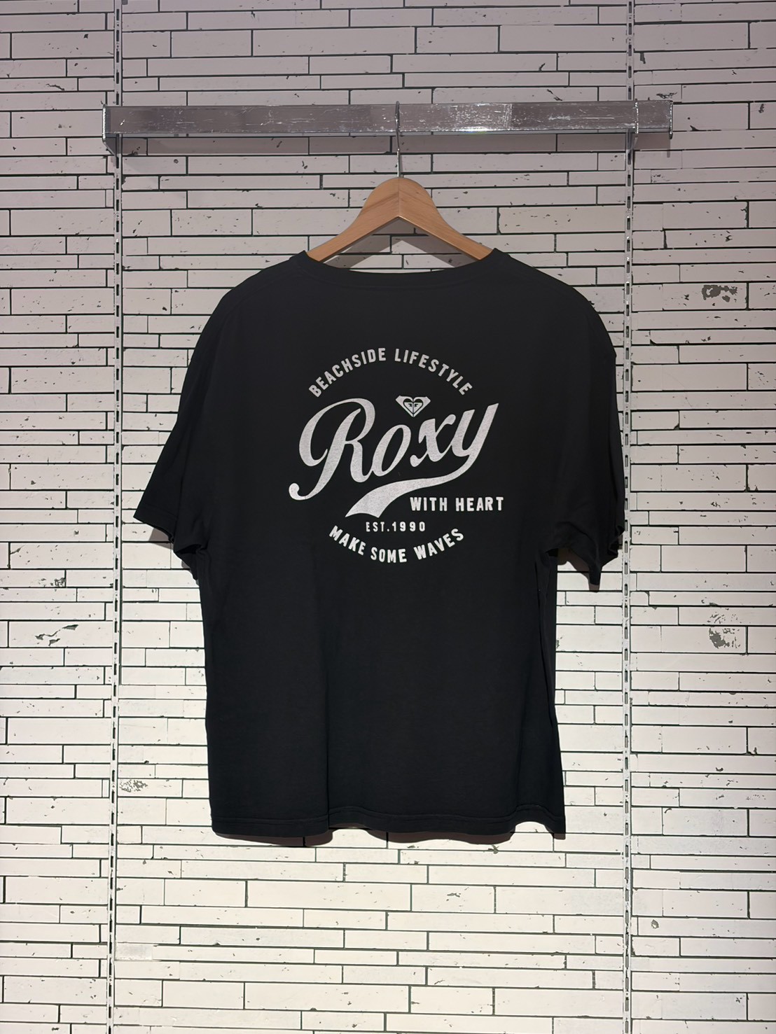 Black T-shirt on a wooden hanger against a white brick wall, featuring a large white 'Roxy' logo and beachwear slogan.