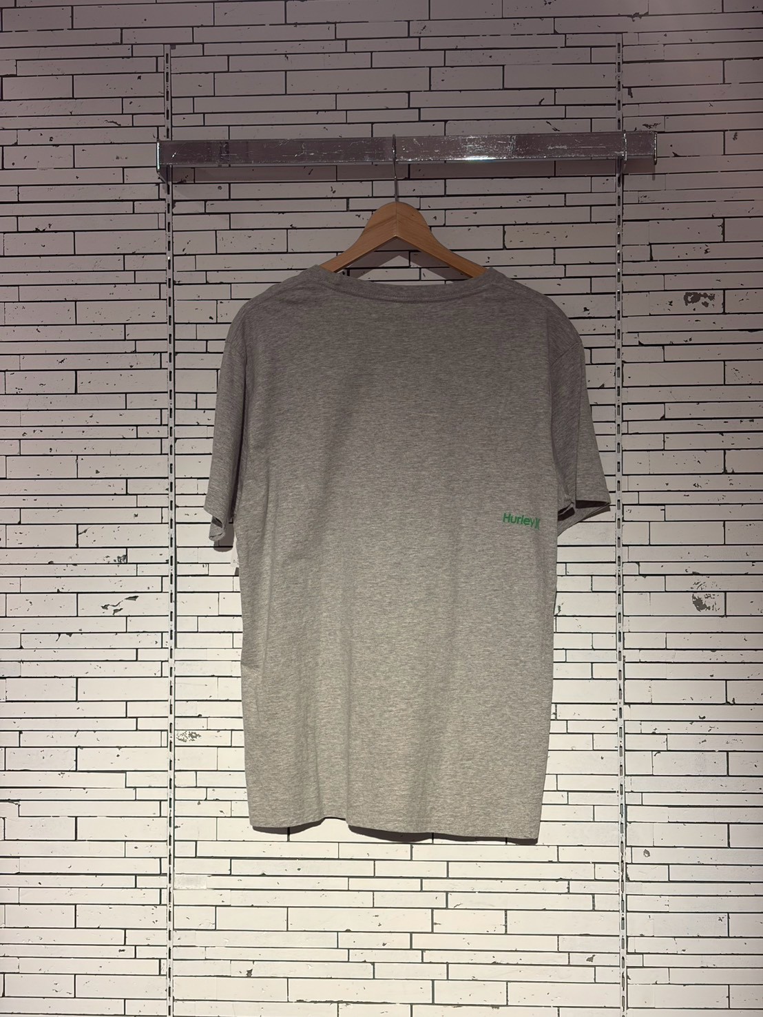 Grey short-sleeve t-shirt on a wooden hanger against a white brick wall, with a small green Hurley logo on the front.