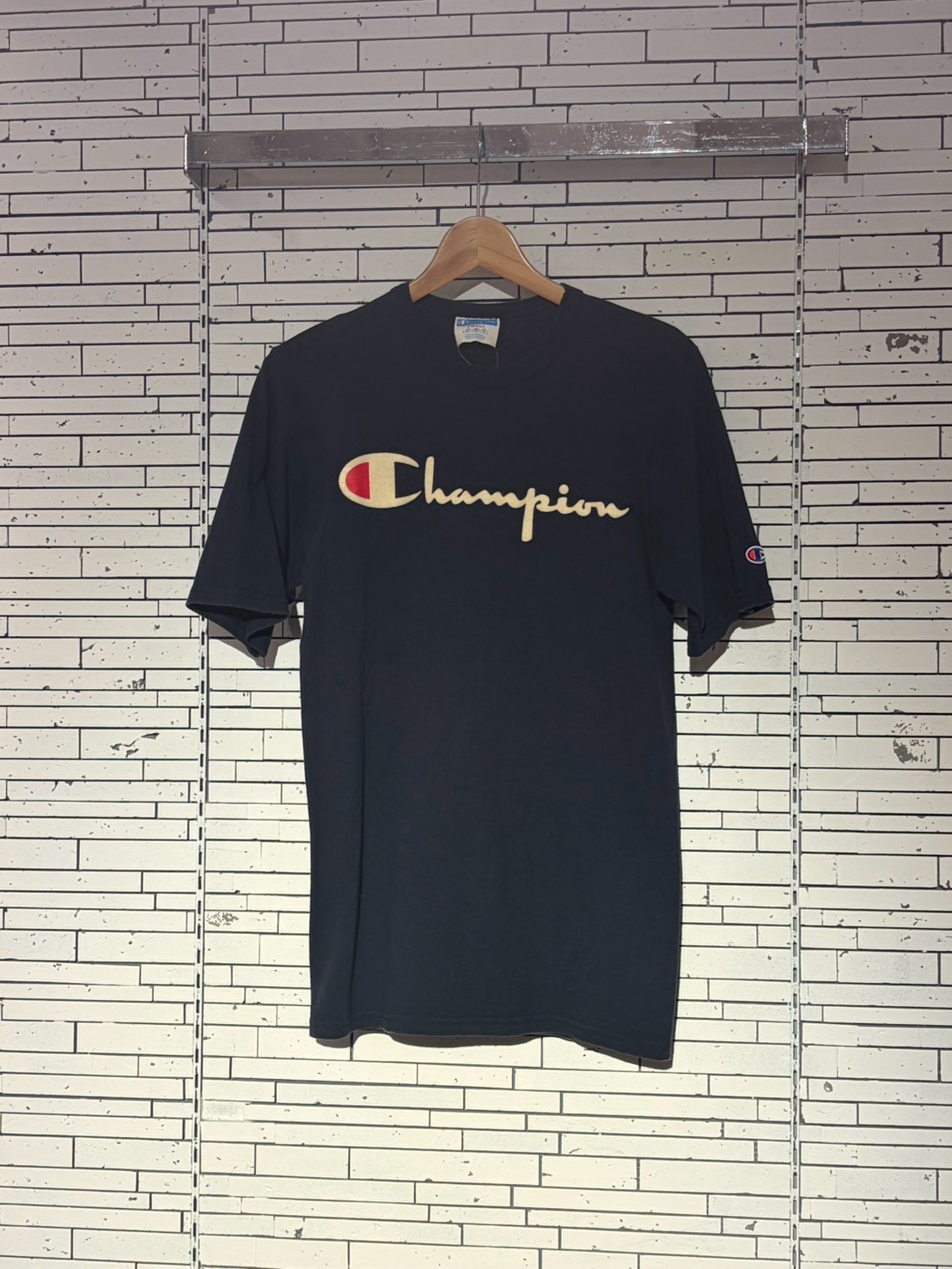 Navy Champion T-shirt hanging on a wooden hanger against a white brick wall background.