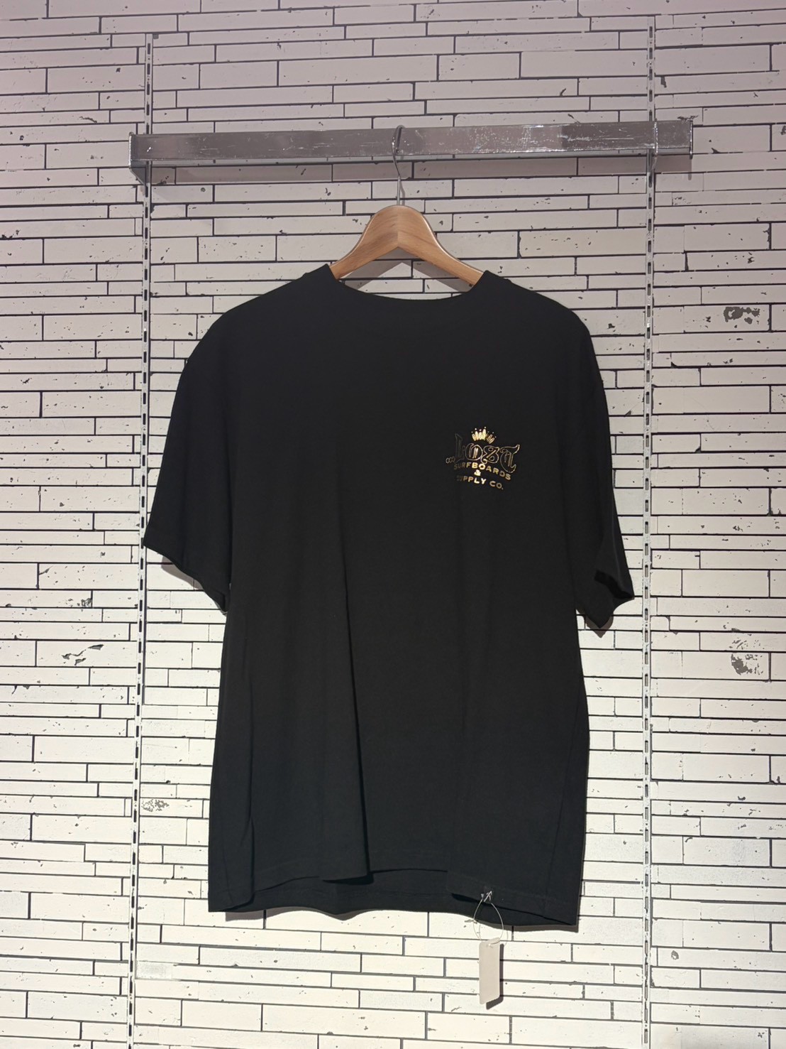 Black short-sleeve T-shirt on a wooden hanger against a white brick wall; small gold logo on the left chest.