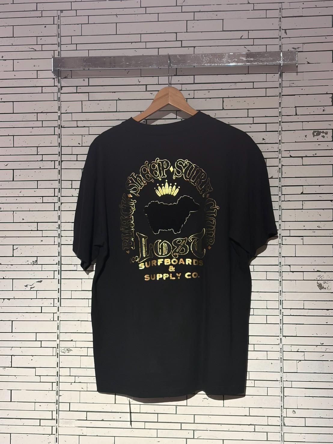 Black t-shirt on a wooden hanger with a gold foil graphic: crown above a dark animal silhouette, surrounded by circular gold text reading a brand name and 'Surfboards & Supply Co.'