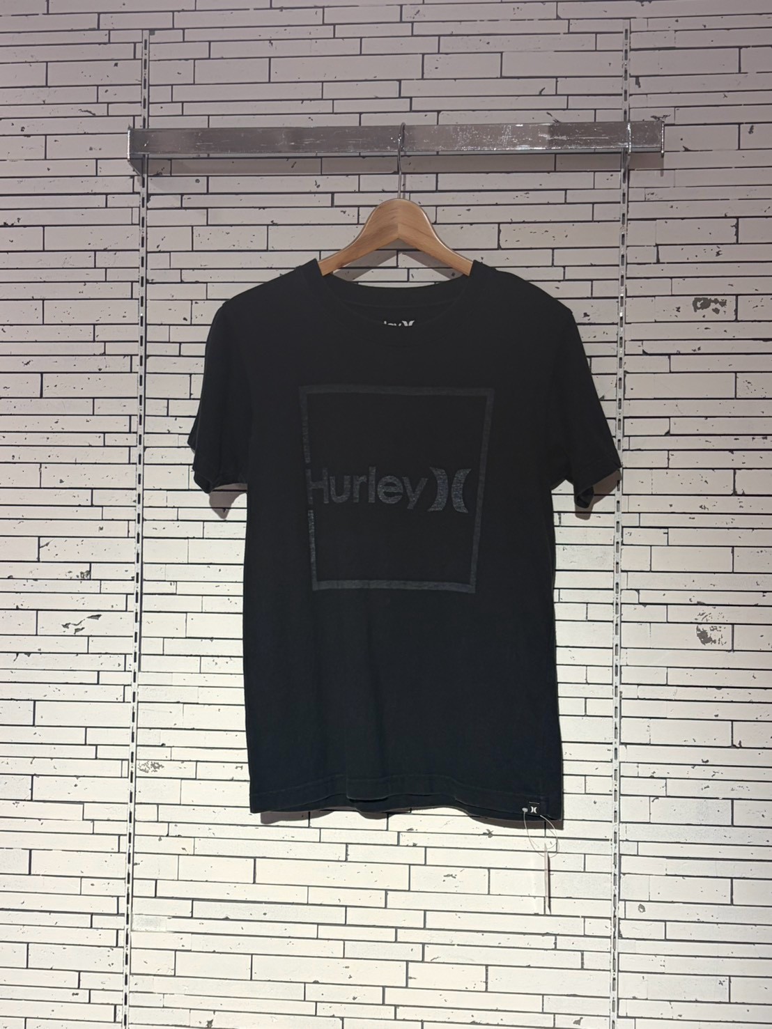 Black Hurley T-shirt hanging on a wooden hanger in a retail display against a white brick wall, front view.