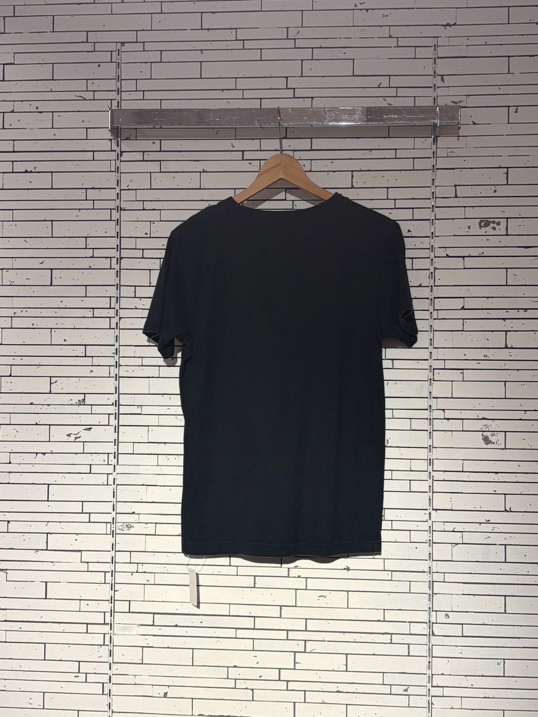 Black t-shirt on a wooden hanger against a white brick wall, viewed from the back.