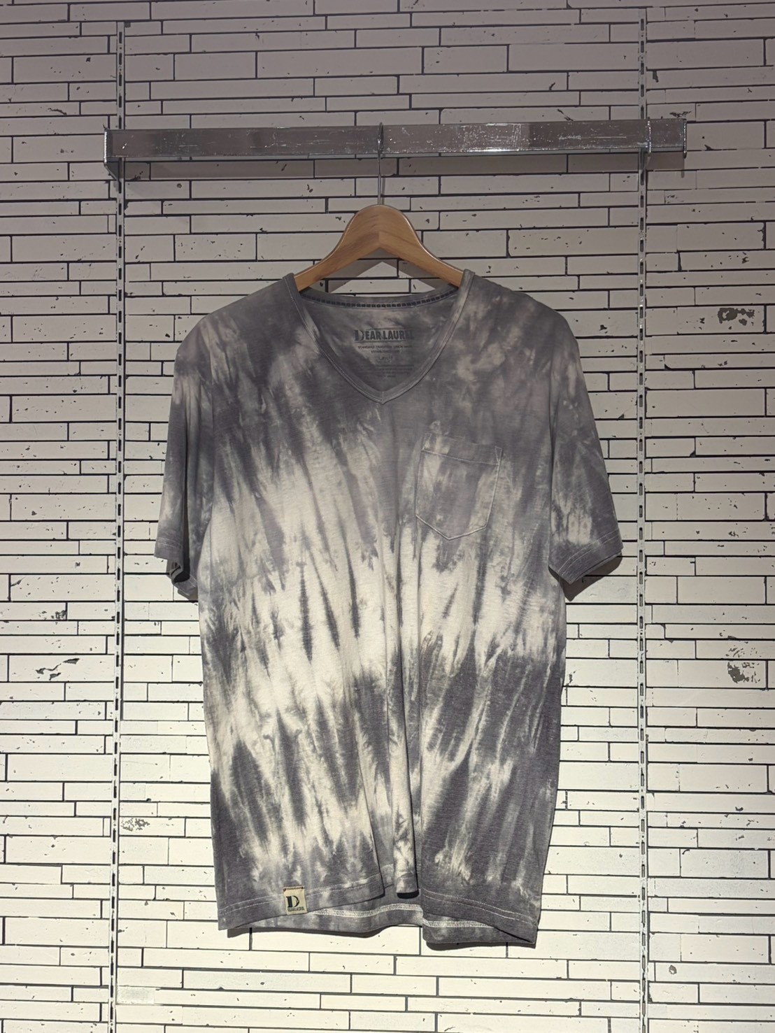 Gray tie-dye V-neck T-shirt with a chest pocket on a wooden hanger against a light brick/tile wall.