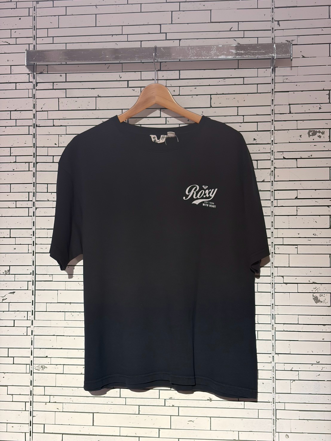 Black T-shirt on a wooden hanger against a white brick wall, featuring a small white Roxy logo on the left chest.