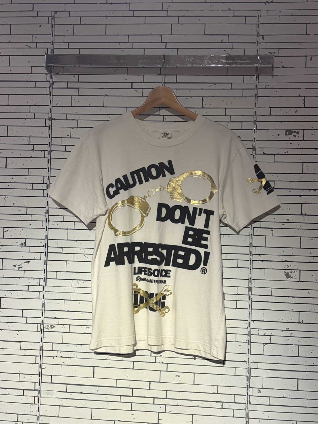 White graphic T-shirt on a hanger with bold black text 'CAUTION DON'T BE ARRESTED!' and gold ring accents against a white wall.