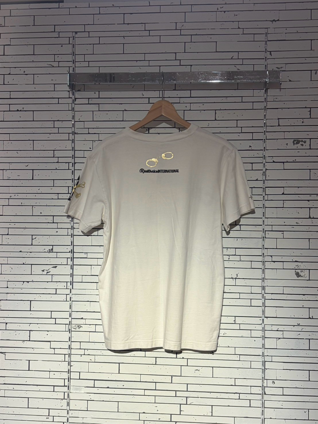 White T-shirt on a wooden hanger against a white brick wall, with a small gold logo on the left sleeve.