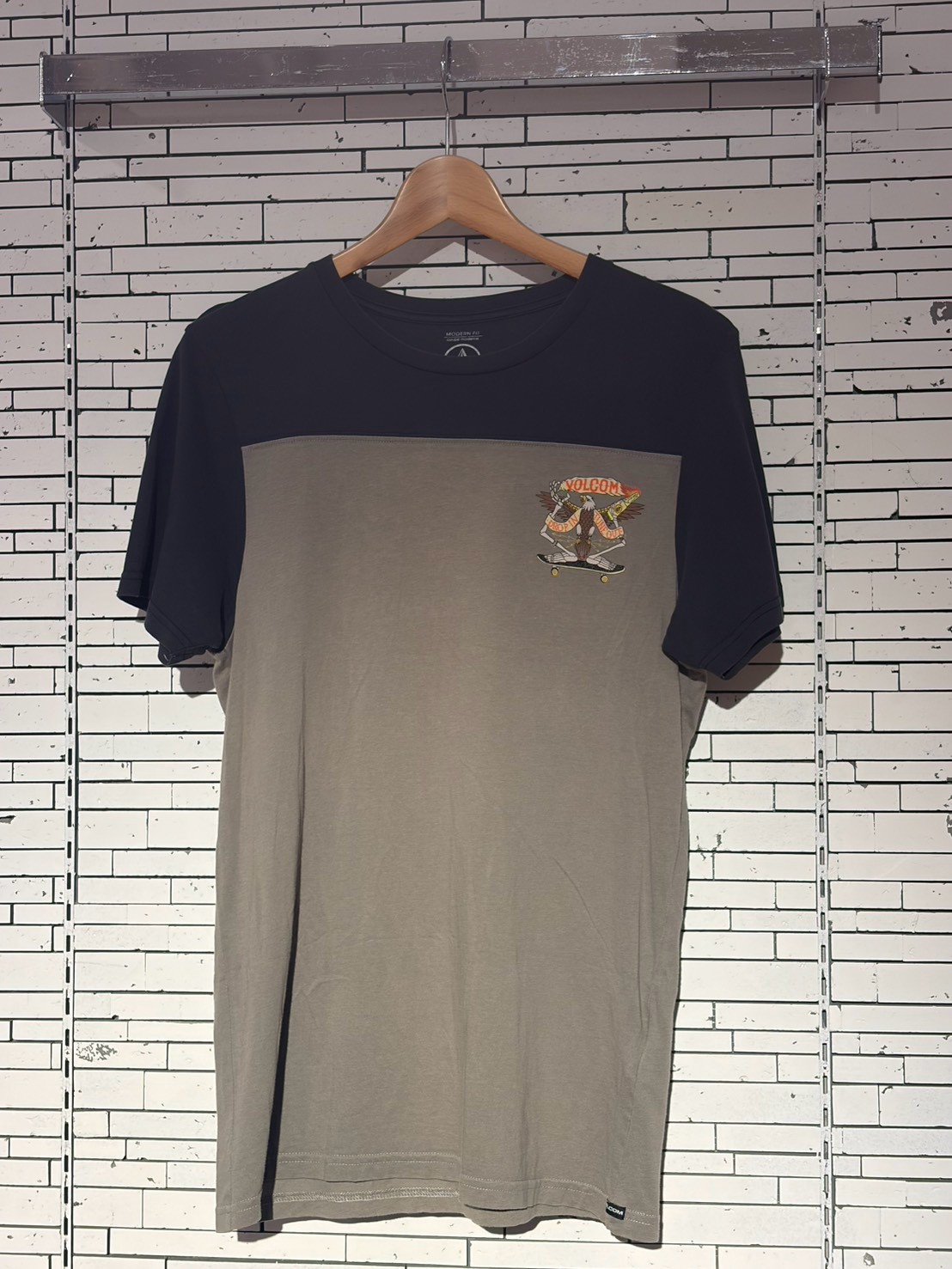 Two-tone short-sleeve t-shirt on a wooden hanger against a white brick wall; small graphic logo on the chest panel.