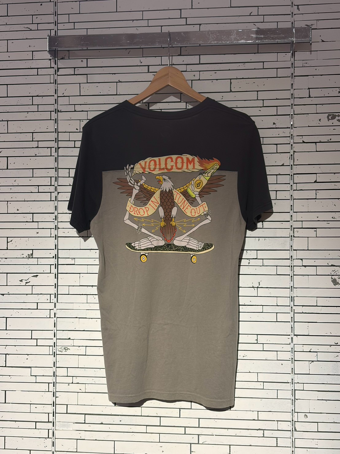 Volcom graphic tee on a wooden hanger against a white brick wall, featuring an eagle with a skateboard and the banner 'Drop In Tune Out'.