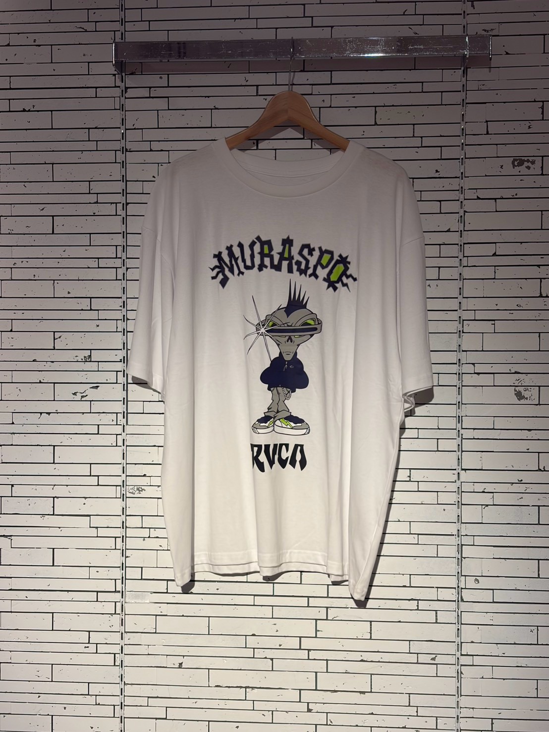 White oversized T-shirt on a hanger featuring a large cartoon alien graphic wearing a hoodie and sneakers, with stylized text across the chest and a brand name at the bottom.