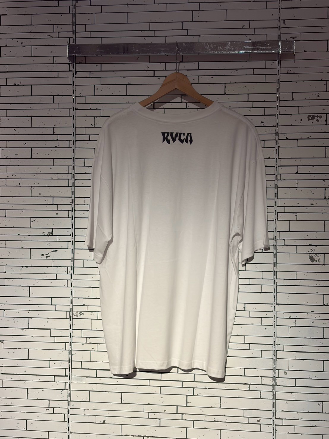 White T-shirt on a wooden hanger hanging against a white tiled wall; RVCA logo visible on the back.