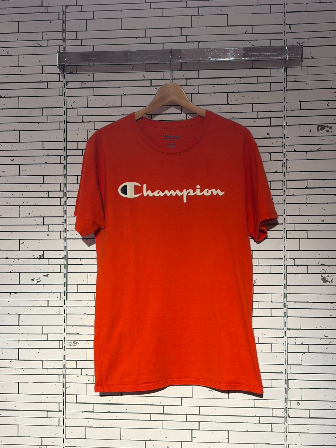 Orange Champion T-shirt hanging on a wooden hanger against a white tiled wall, front view.