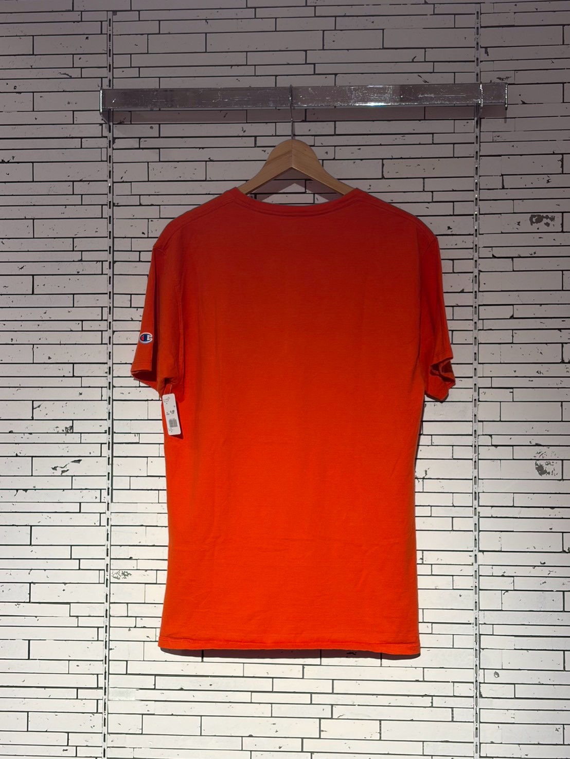 Bright orange short-sleeve T-shirt on a wooden hanger against a white brick wall.