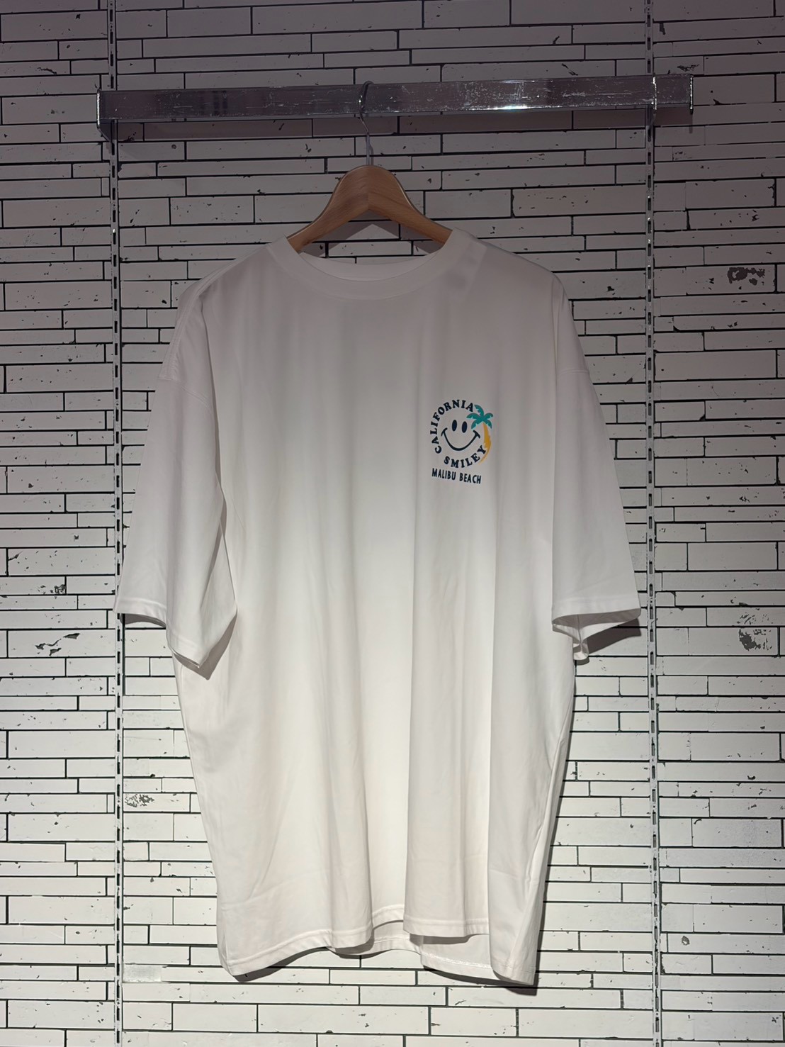 White short-sleeve white T-shirt on a wooden hanger against a light brick wall with a small logo on the left chest reading 'California Smile Malibu Beach' with a palm tree design.