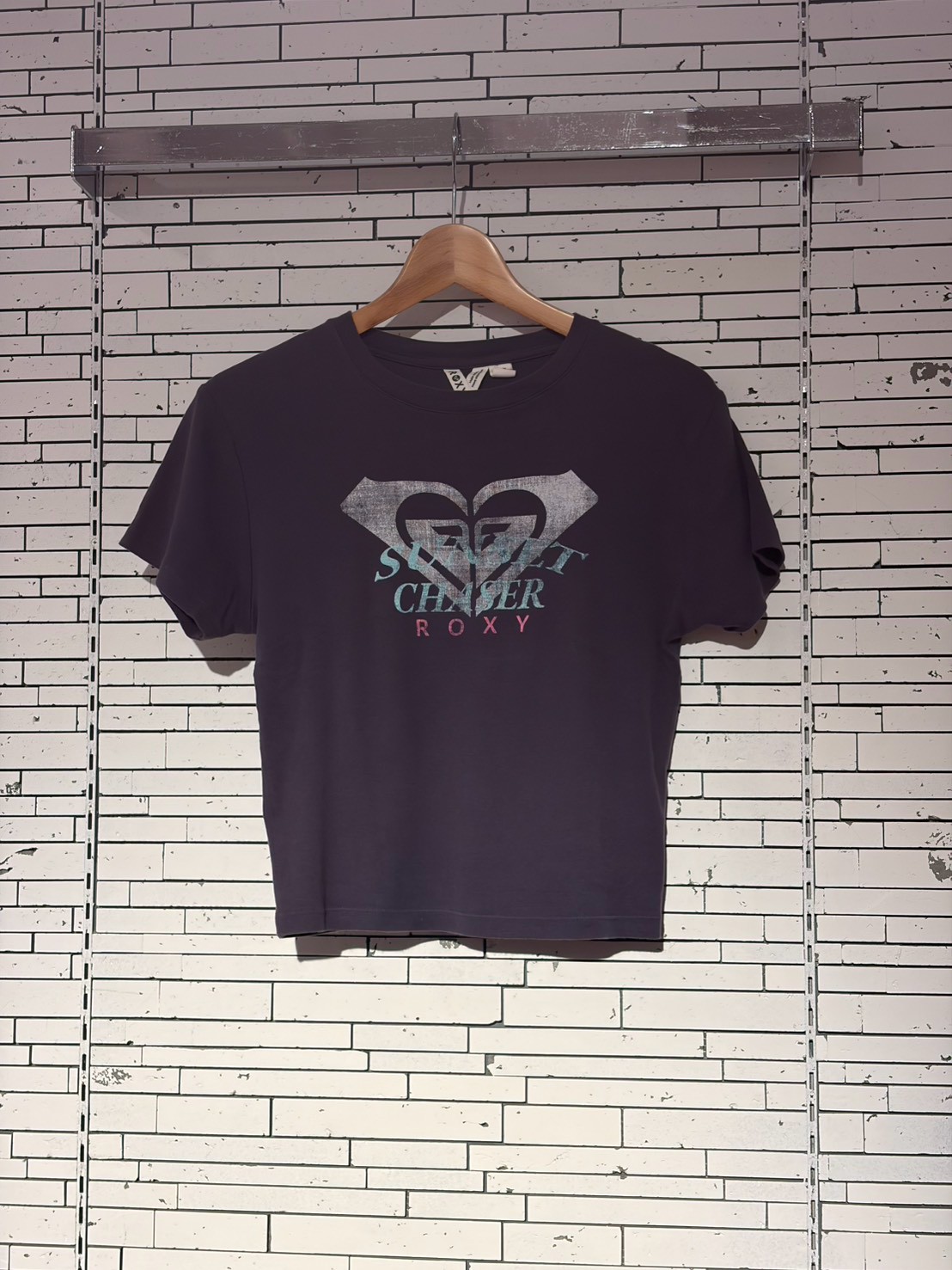 Navy T-shirt on a wooden hanger against a white brick wall, featuring a faded heart-shaped graphic and the word 'ROXY' on the front.