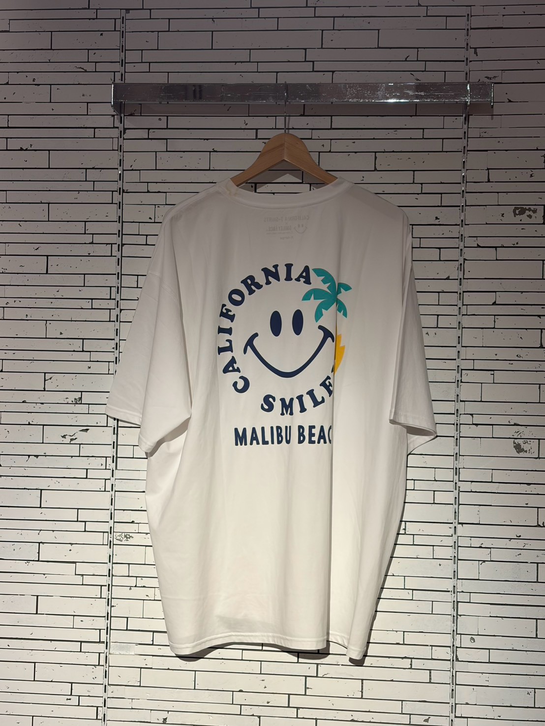 White T-shirt on a hanger with a California Smile Malibu Beach graphic (smiley face, palm tree, sun).