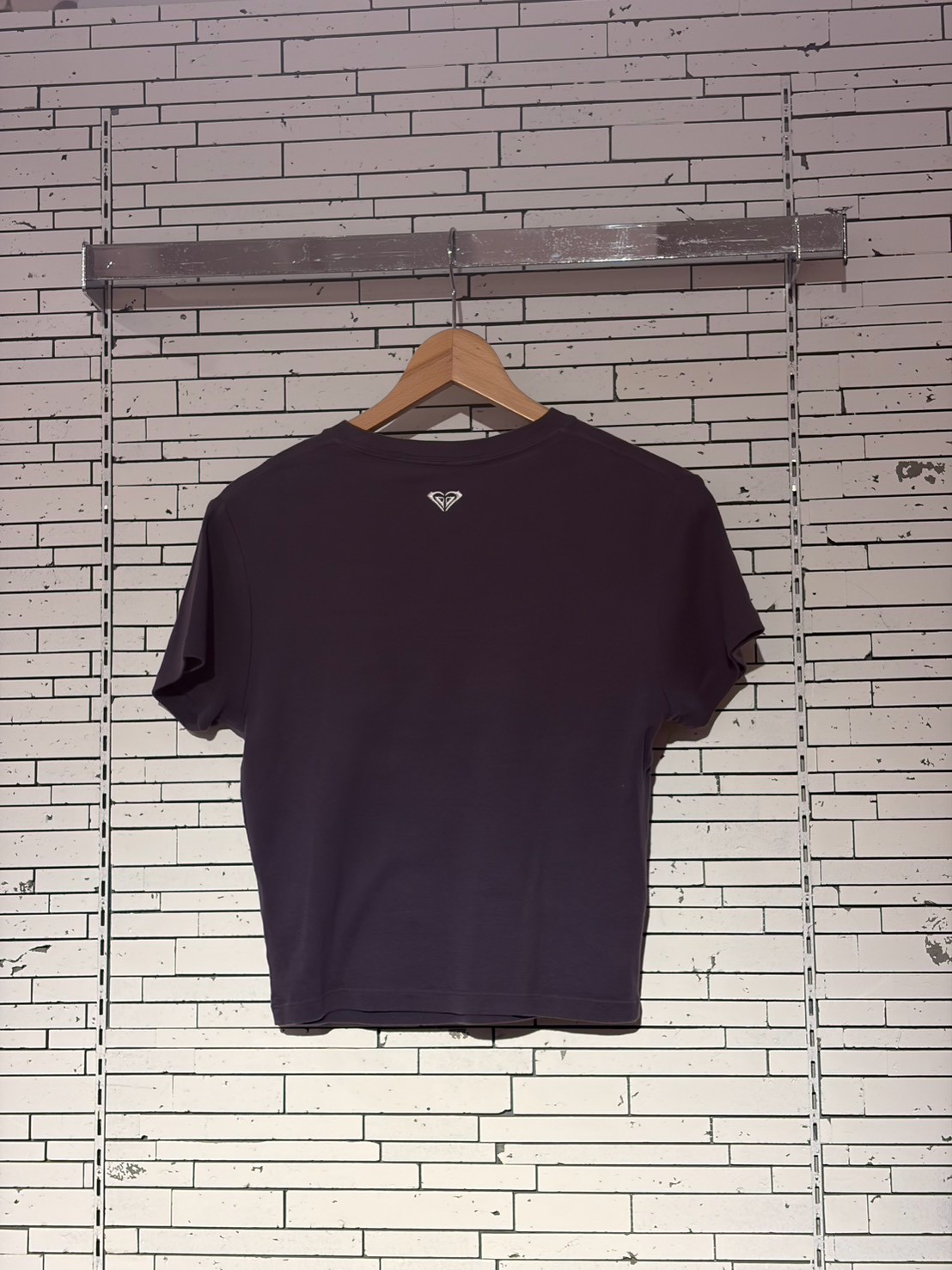 Dark purple T-shirt hanging on a wooden hanger against a white brick wall, small logo near the neck area on the back